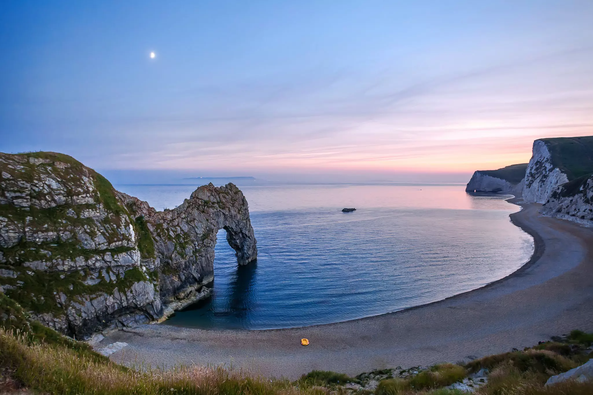 A rock arch stretches into a small bay at twilight. A small yellow boat sits on the shore. Jurassic Coast, England.