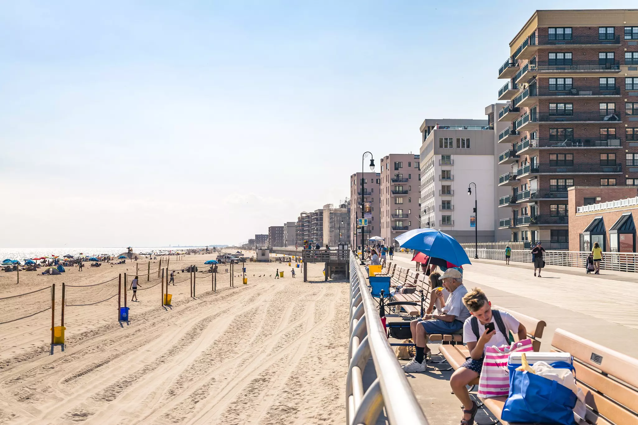 People sitting on benches on a boardwalk by a sandy beach.