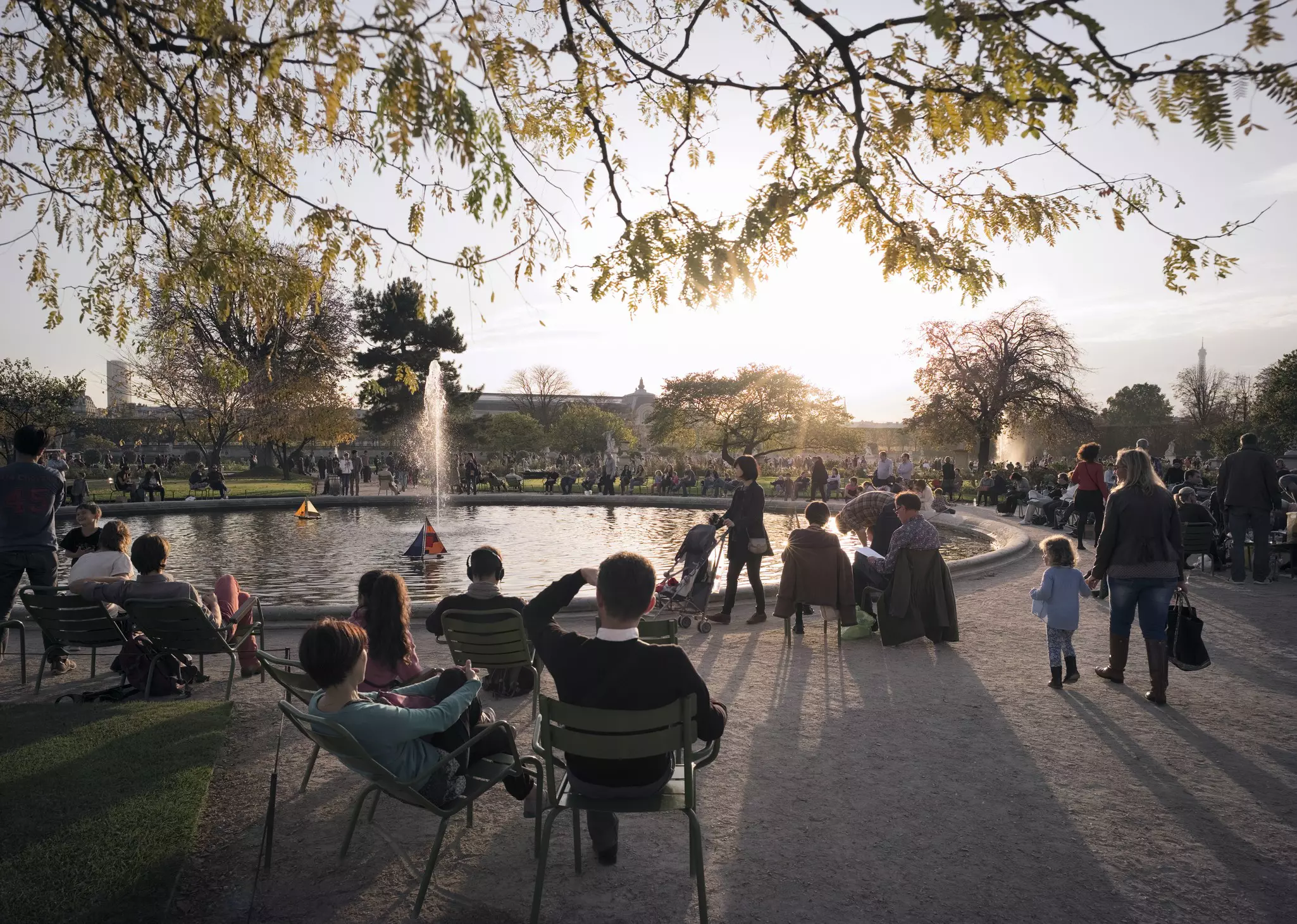 A sunny day in a park. People are out with children playing with toy sailboats on the water, while others sit on deck chairs facing the pond.