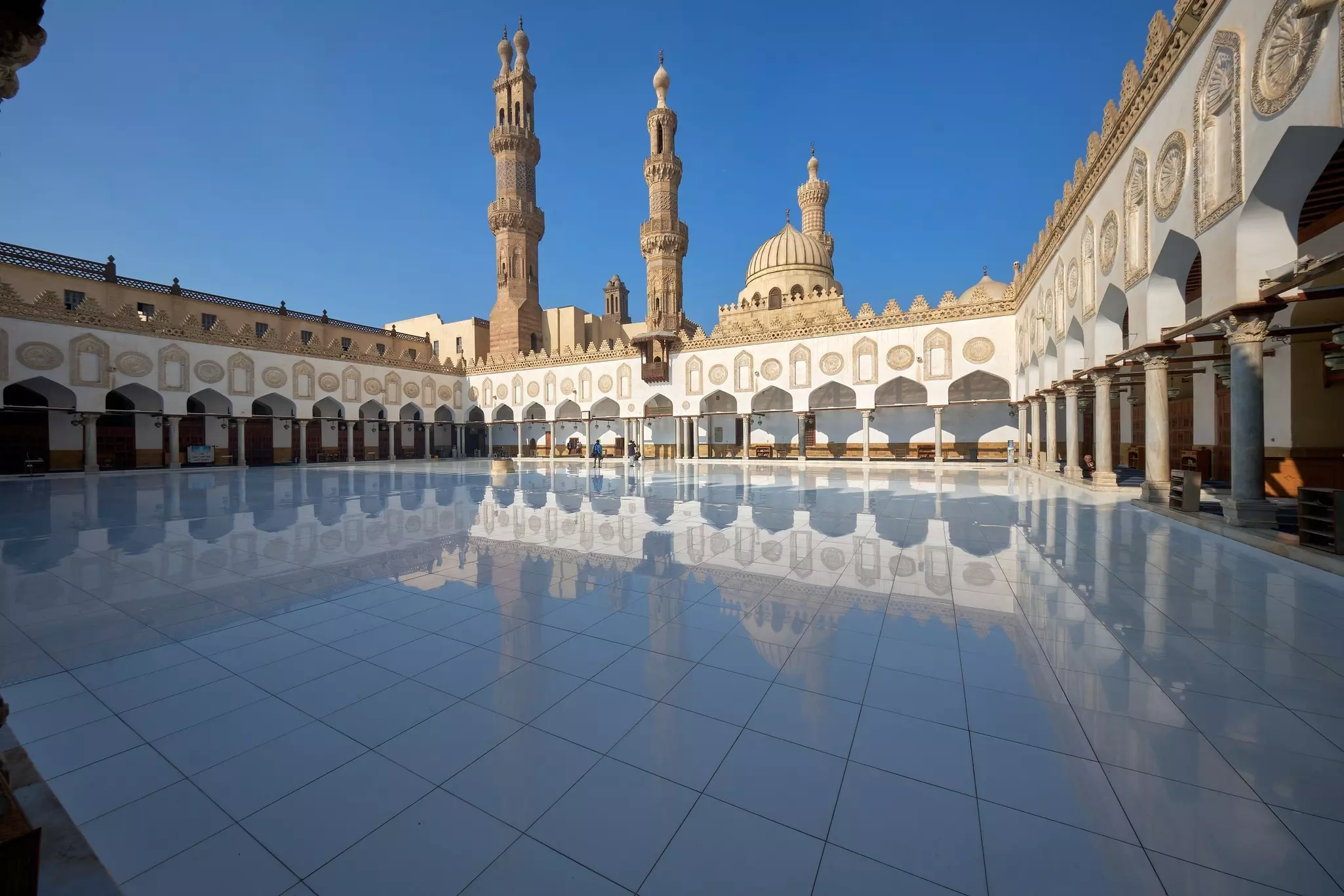 The courtyard of a mosque in Cairo, Egypt, reflects the surrounding arcade and the minaret; the blue sky is cloudless.