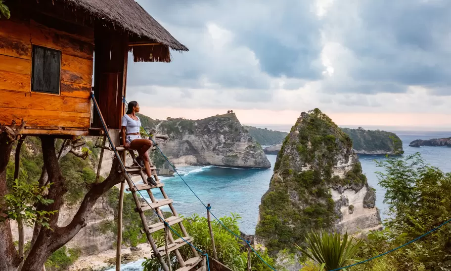 Nusa Penida treehouse with a woman on its ladder above sea stacks, cliffs and turquoise water under cloudy skies
