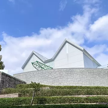 Angular white roofs of a modern museum above a curved stone wall and terraced hedges under a blue sky