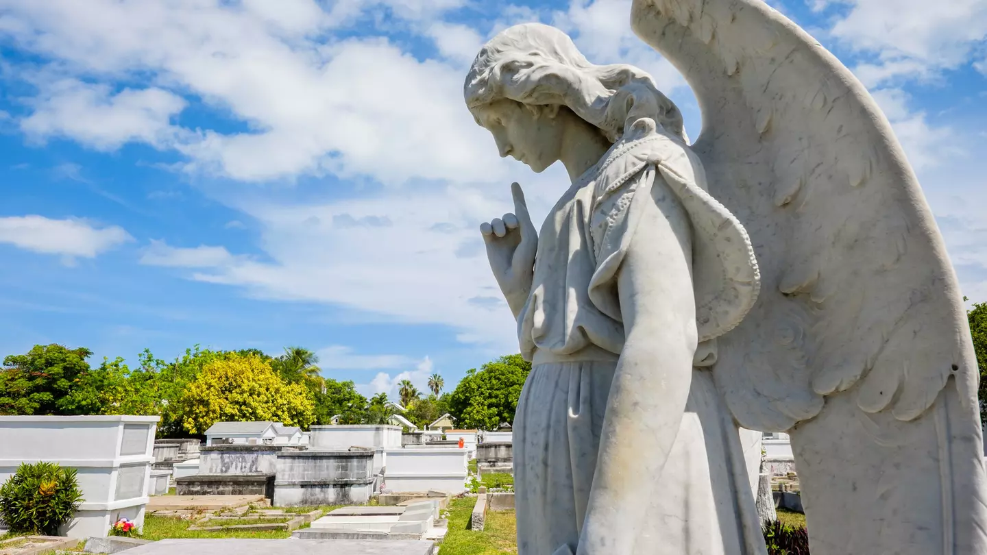 A stone sculpture of a winged angel is in a cemetery with mausoleums.