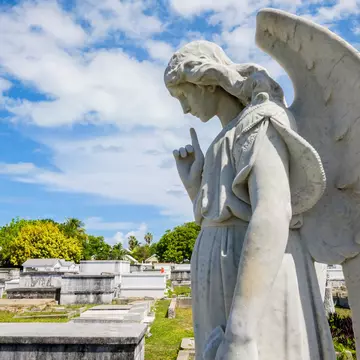 A stone sculpture of a winged angel is in a cemetery with mausoleums.