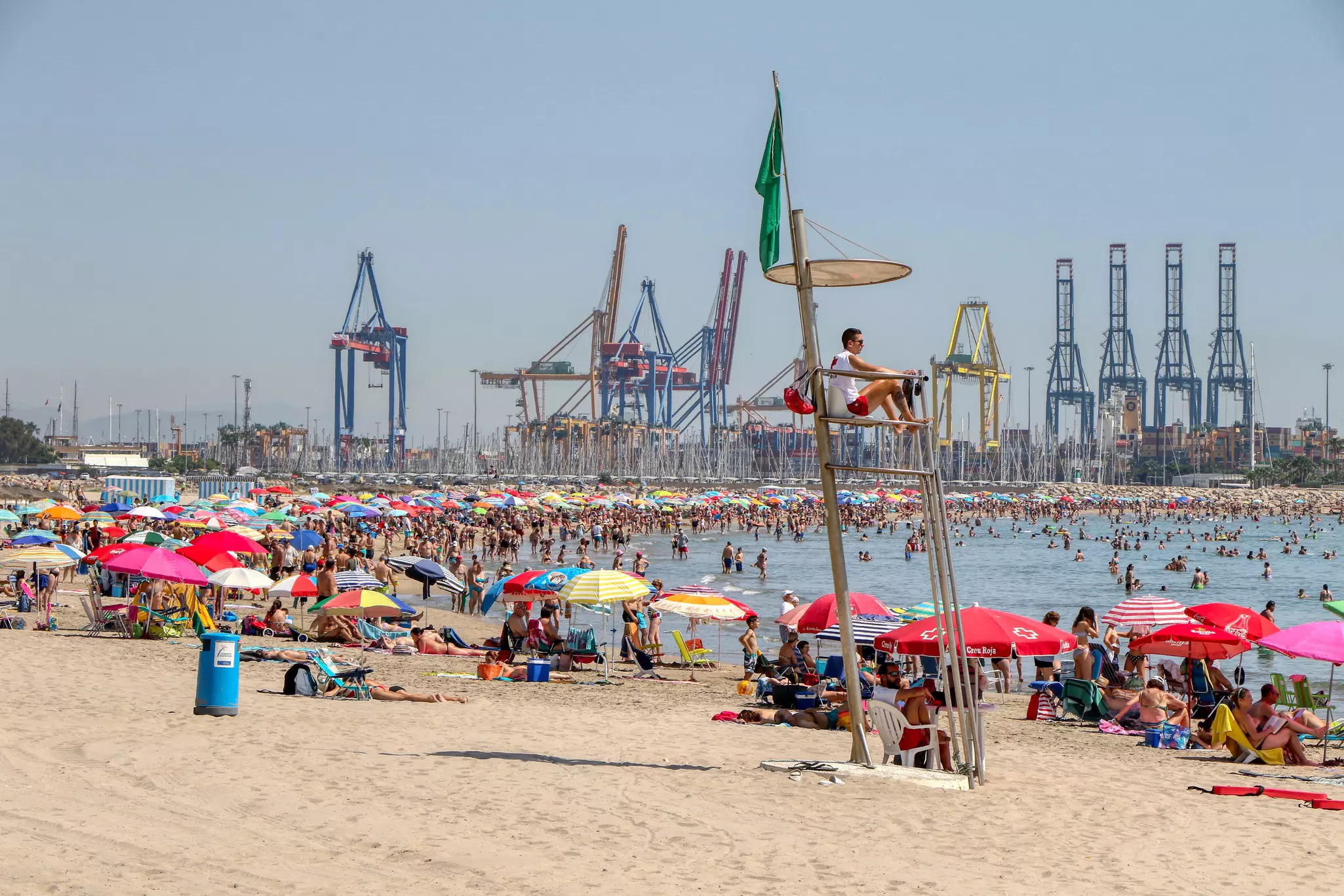 A busy sandy beach with people under sunshades. A port is in the distance.