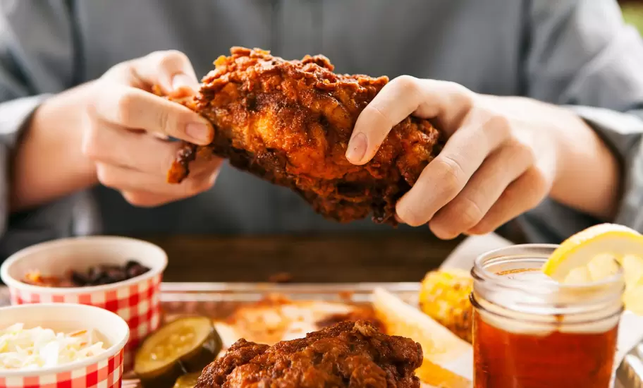 Hands lift a piece of Nashville hot chicken over a tray with pickles, coleslaw, beans, corn and a jar of iced tea