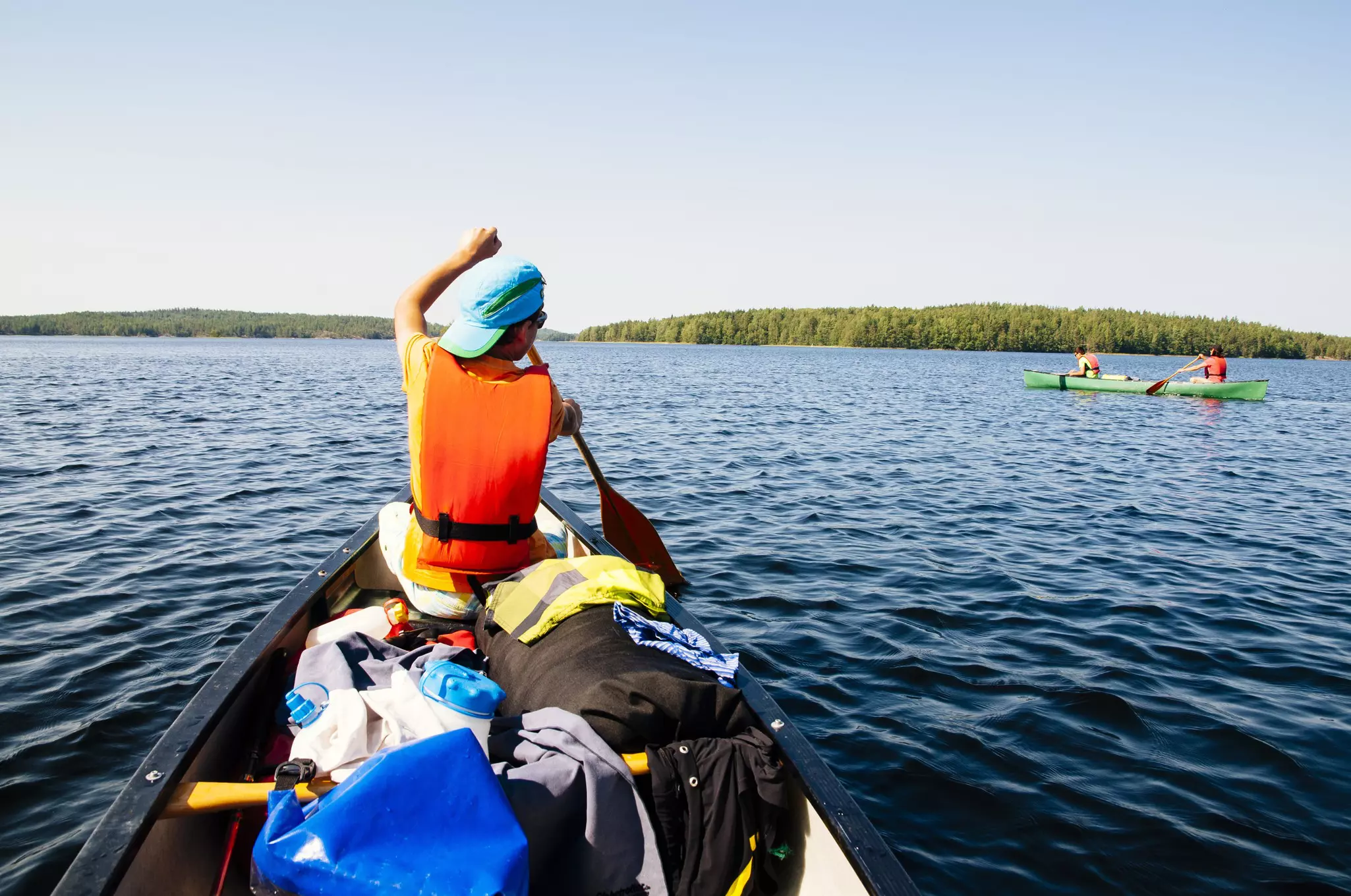 A young man in an orange life vest and blue hat paddles a canoe full of equipment. Another couple paddles next to him.