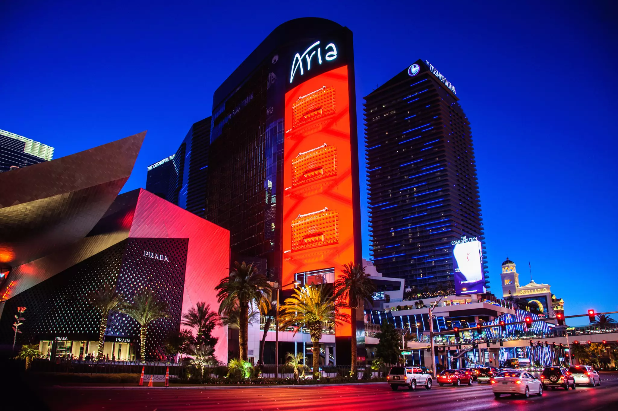The Aria hotel partially illuminated in red with palm trees in front, with the Cosmopolitan hotel striped in blue nearby