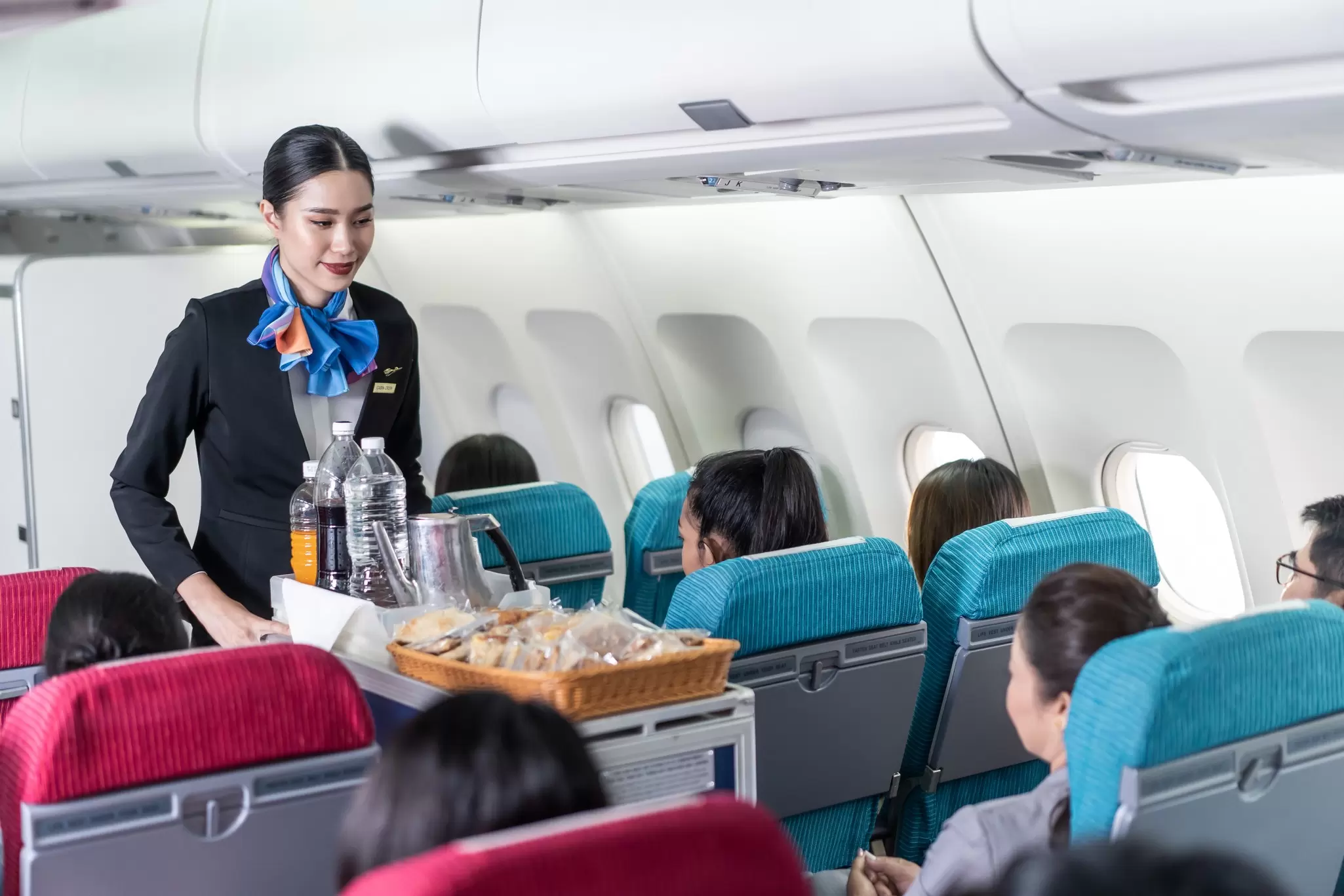 Flight attendant in a blue scarf pushes a cart with water bottles and snacks along an airplane aisle past seated passengers
