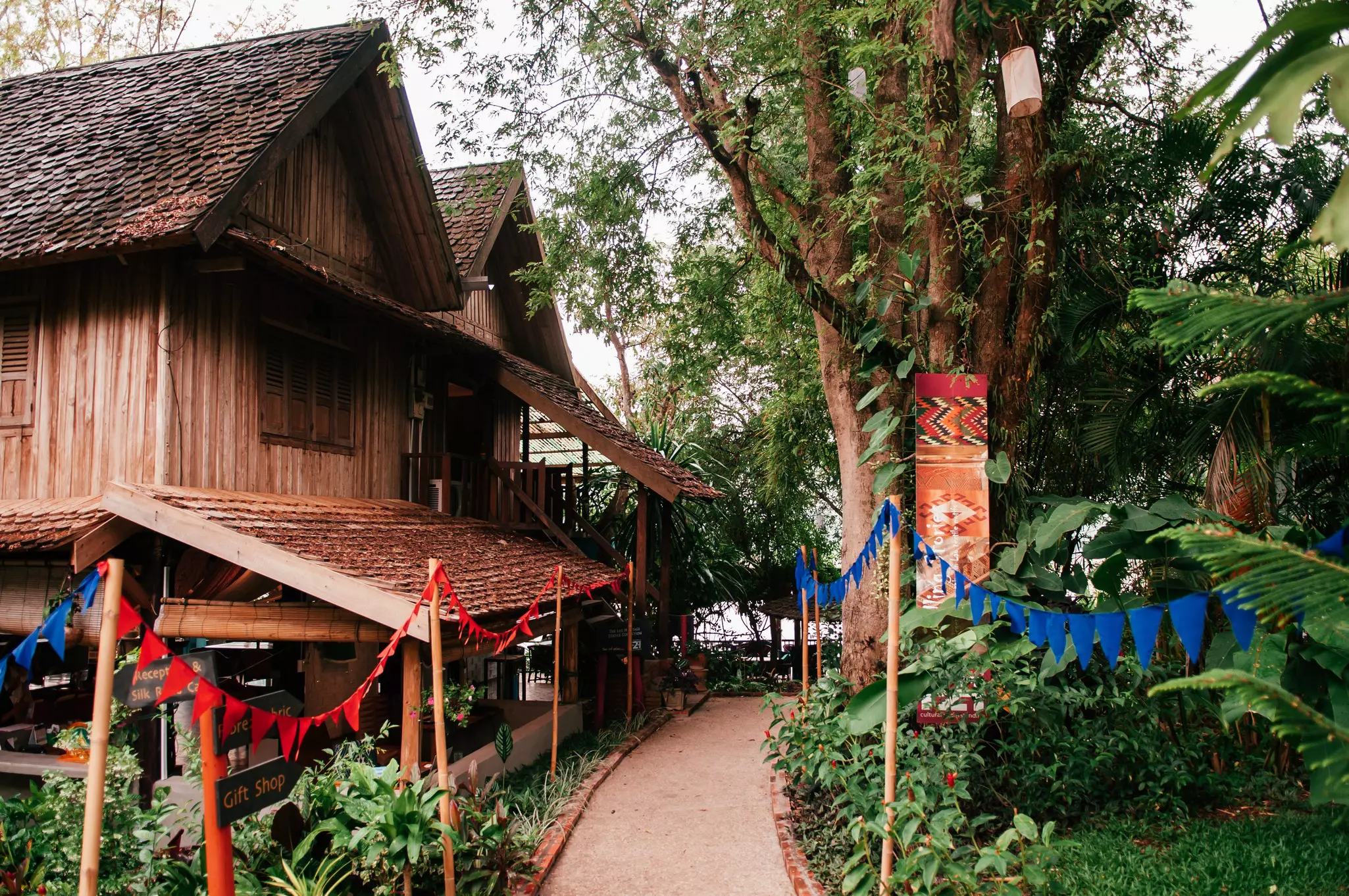 A walkway lined with red and blue flags and greenery. A wooden house is to its left, and mostly greenery and trees are on its right.