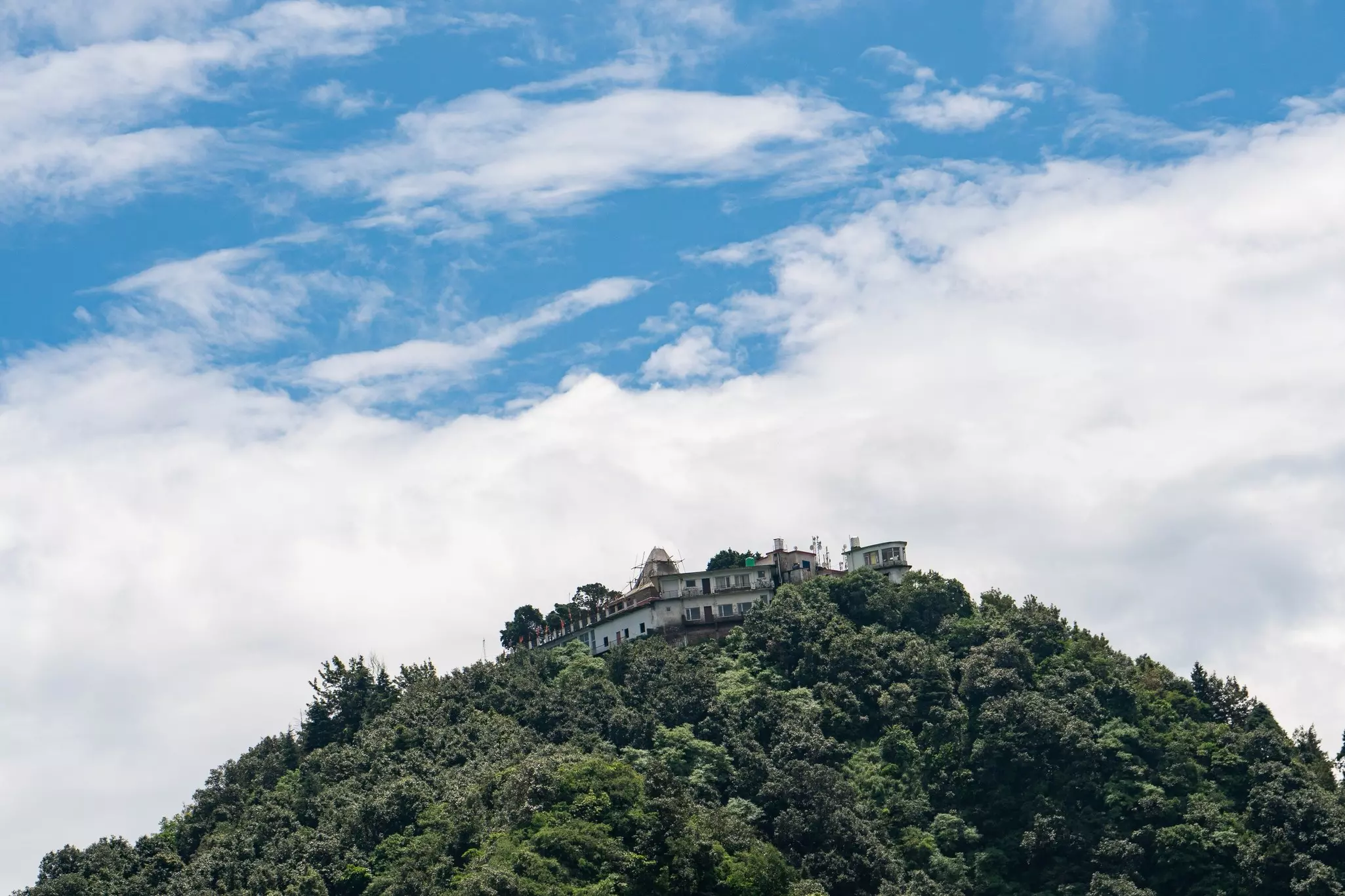 Temple on a forested mountain surrounded by a blue and cloud-filled sky.