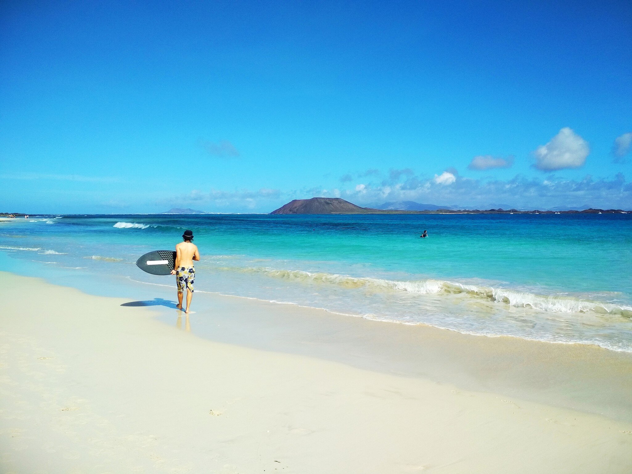Man carrying a surfboard along pale sand beside turquoise surf, a low brown volcanic island on the horizon.