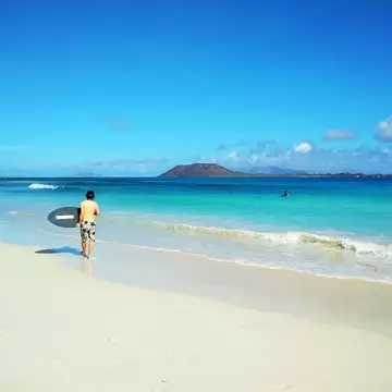 Man carrying a surfboard along pale sand beside turquoise surf, a low brown volcanic island on the horizon.