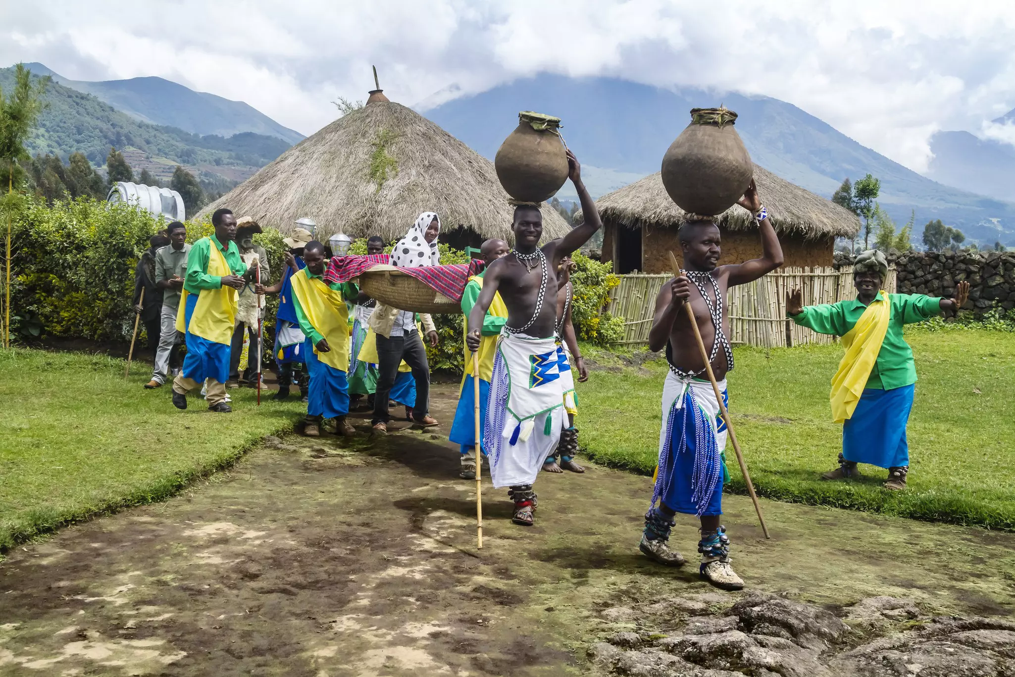 A kneeling Batwa man blows on a collection of dry grass in his hands that smokes; to his left is a boy holding the stick that was used to create the initial spark by spinning.