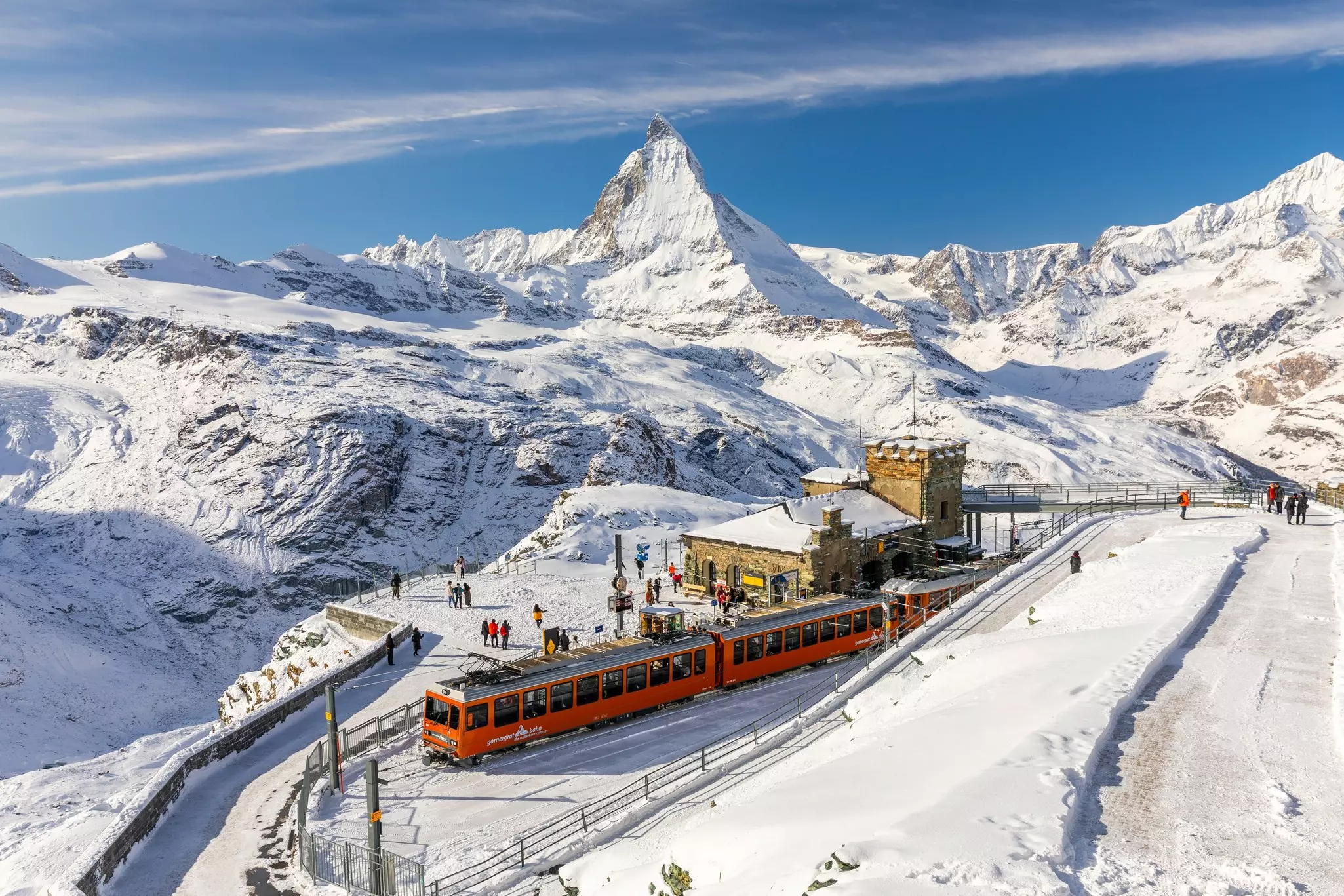 A small red train stopped at an alpine station with a distinctive pointed mountain peak towering over it.