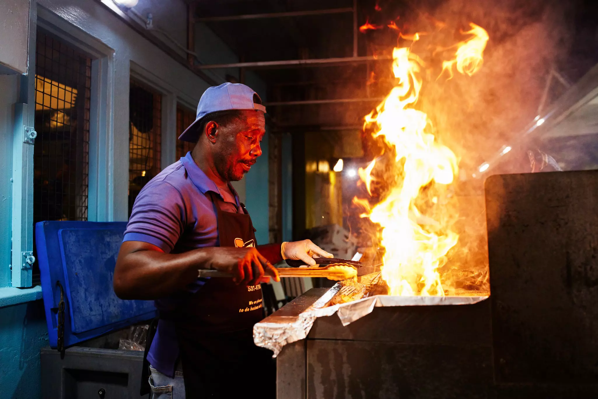 A man stands at a grill in the evening as flames shoot up.