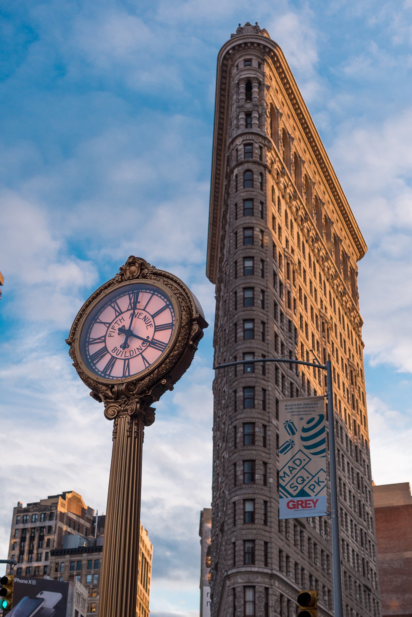 Narrow wedge-shaped skyscraper lit by warm sun behind an ornate street clock reading "Fifth Avenue Building" under blue sky