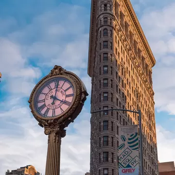 Narrow wedge-shaped skyscraper lit by warm sun behind an ornate street clock reading "Fifth Avenue Building" under blue sky