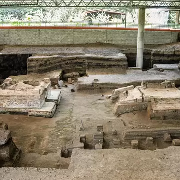 Excavated earthen house platforms and low walls in a sunken pit beneath a steel protective roof.