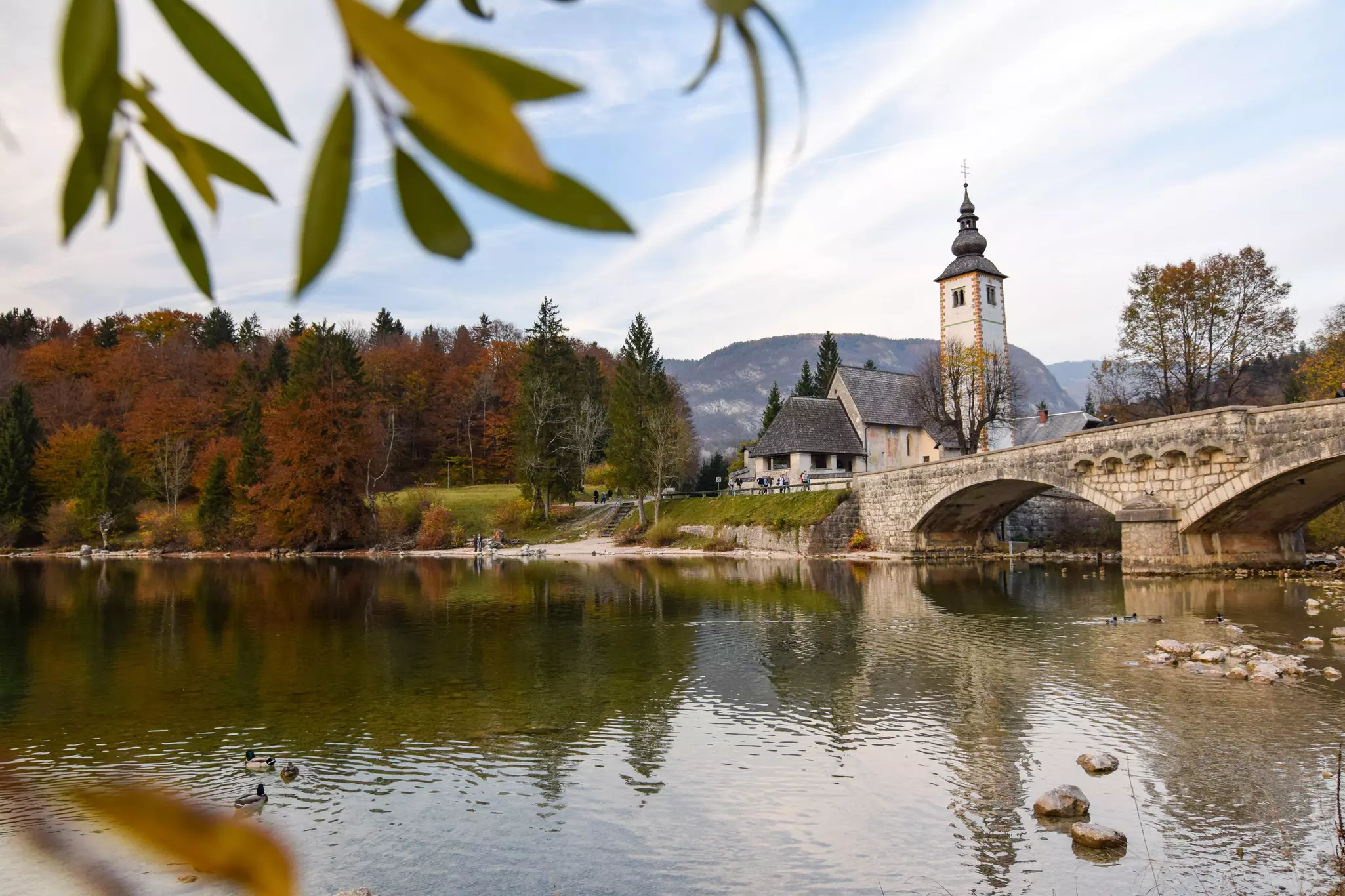 A church sits behind a lake with an arched bridge passing over the lake. Mountains and fall foliage in the background