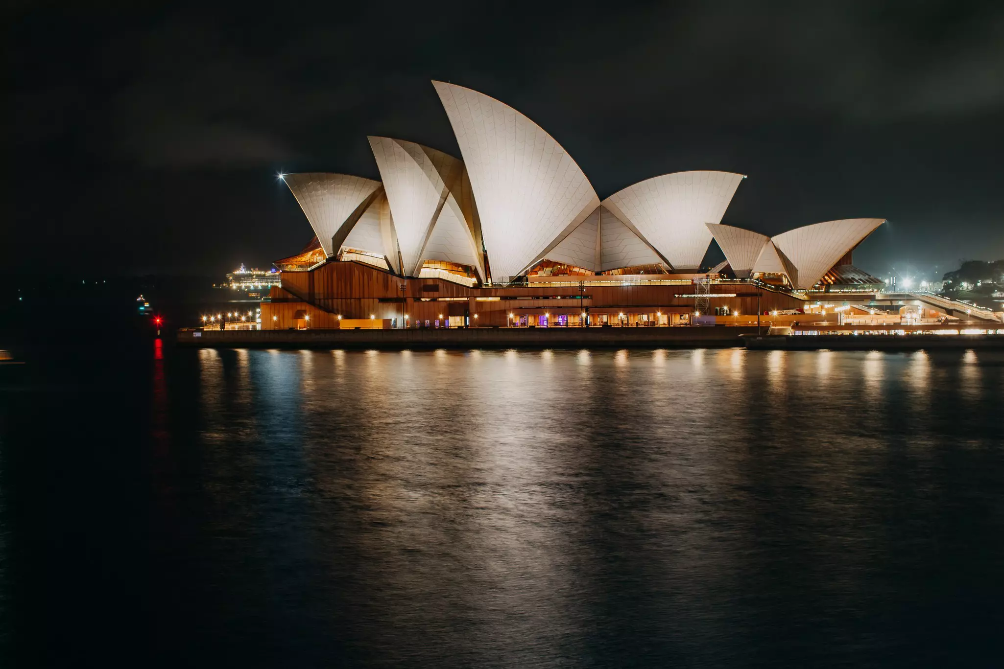 Wide shot of iconic Sydney Opera House with its multiple sail-shaped roofs, a curved harbor bridge in the distance and the harbor in the foreground at night.