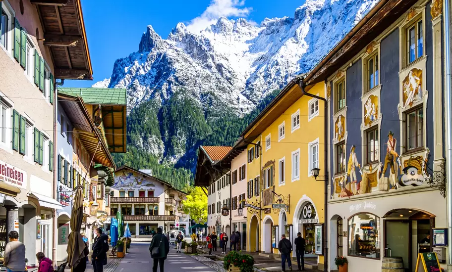Mittenwald street of frescoed Bavarian houses with shops and strolling people, snowy Karwendel peaks rising behind