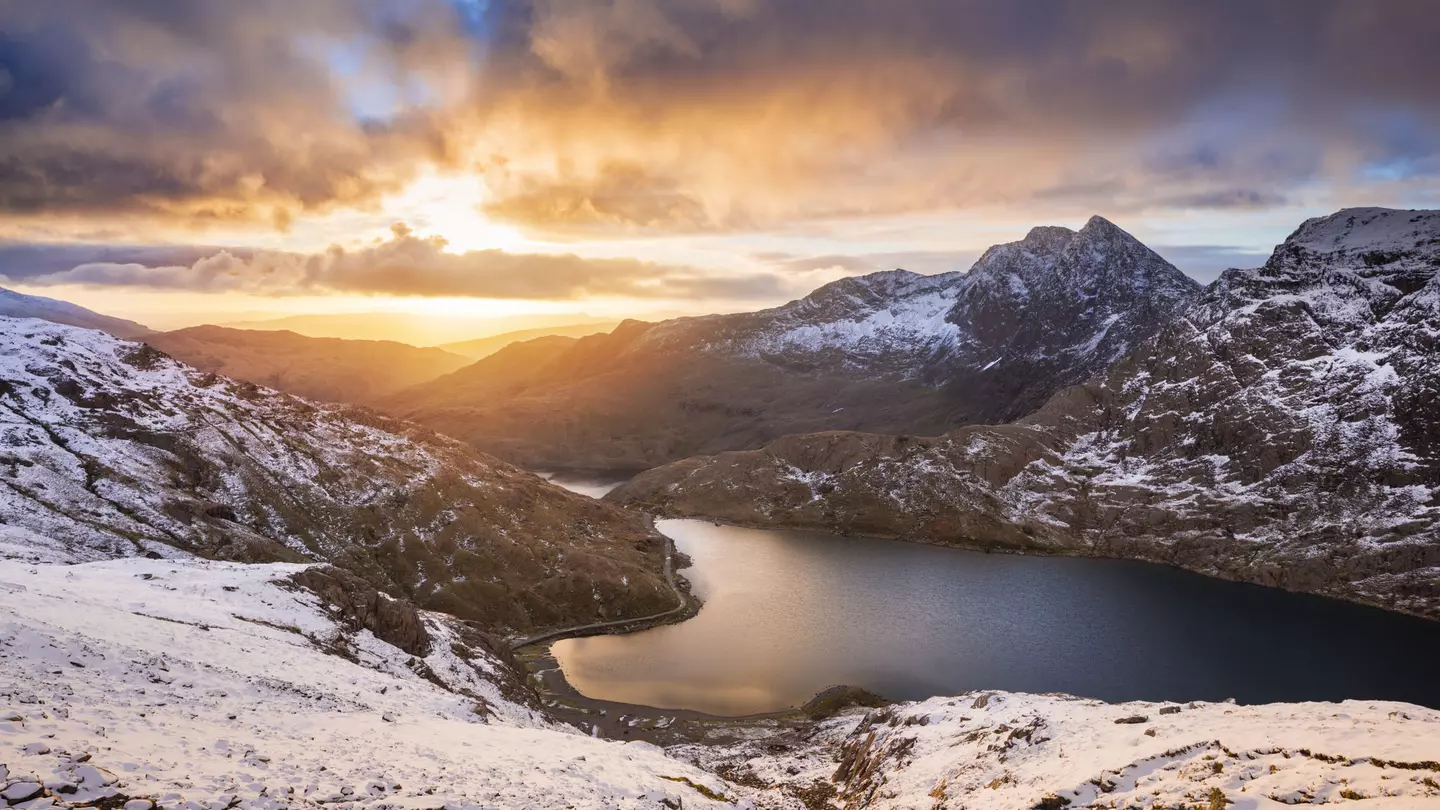 The sun rises over a lake in a snowy mountain landscape, creating orange streaks that contrast with the grey clouds
