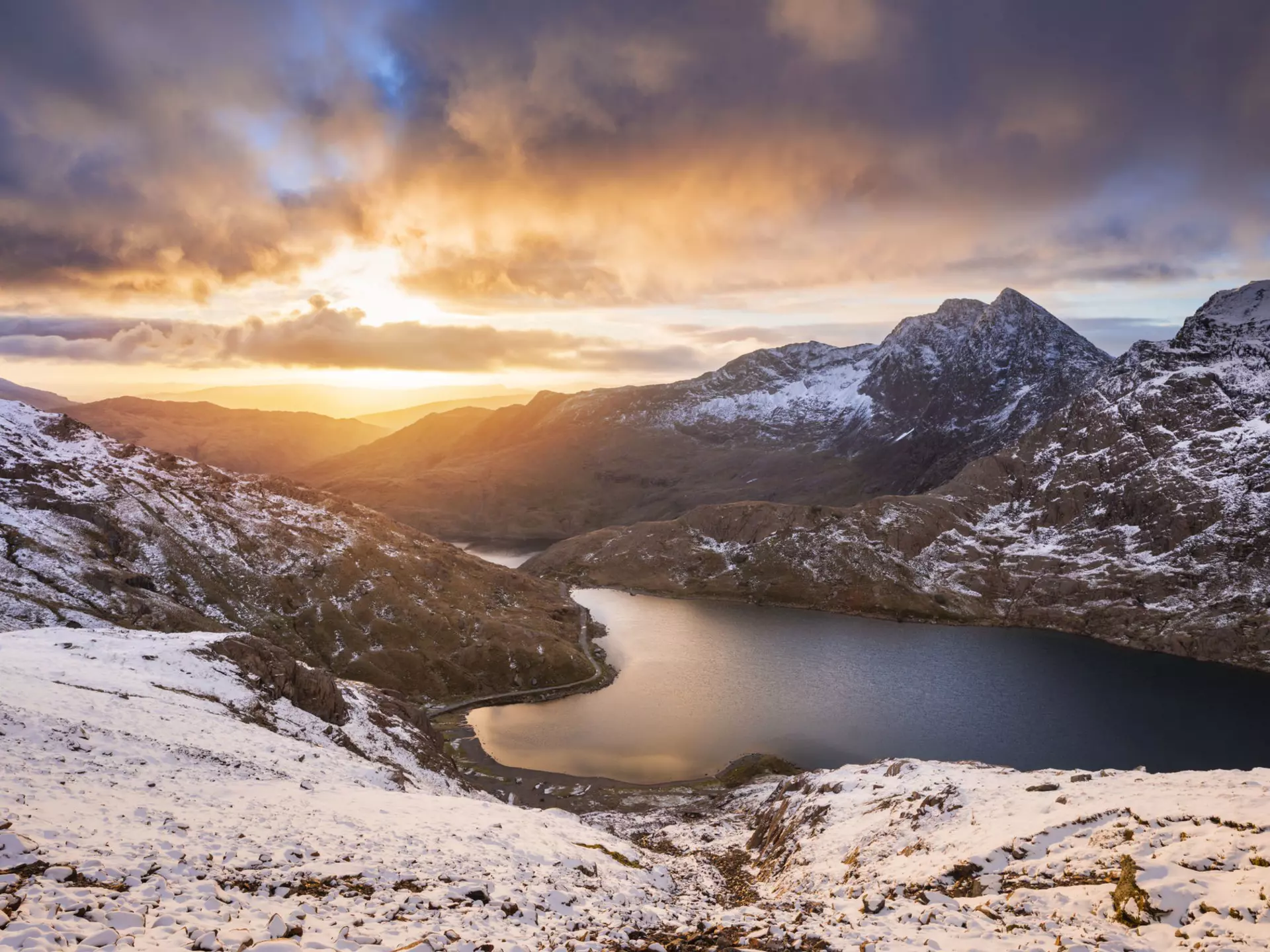 The sun rises over a lake in a snowy mountain landscape, creating orange streaks that contrast with the grey clouds