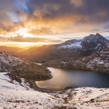 The sun rises over a lake in a snowy mountain landscape, creating orange streaks that contrast with the grey clouds