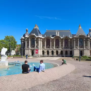 A grand Belle Époque museum with slate roofs and a colonnaded facade, seen across a shallow fountain where people sit on the rim.