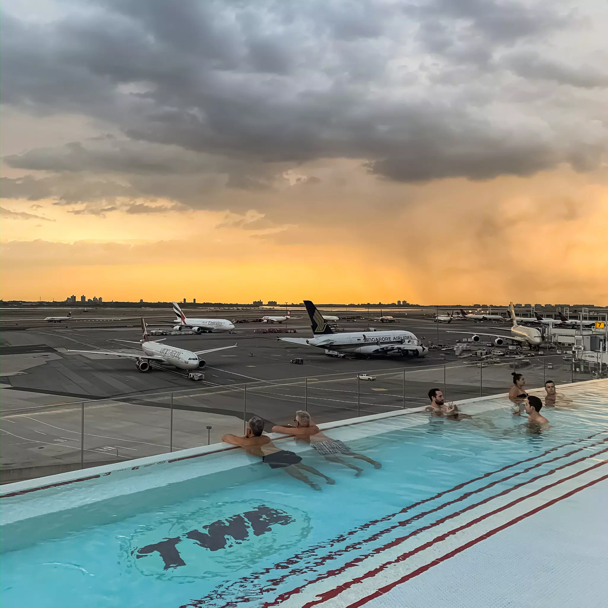 People in a blue swimming pool on roof look toward an airport tarmac with parked planes.
