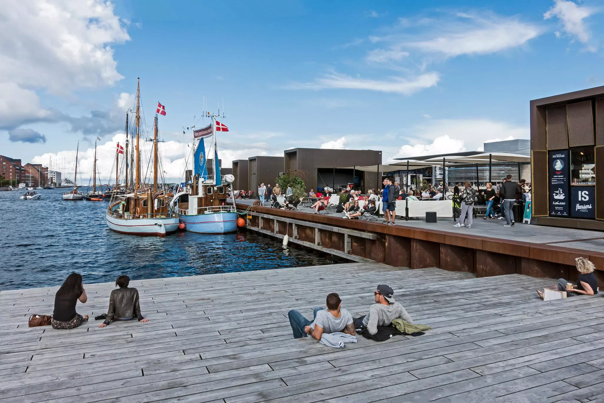 People sitting on a wooden deck facing water in an inlet; there are cafes to one side and boats in the water.