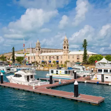 A small marina with boats moored to the boardwalk. A large naval building with two clock towers stands on the shore.