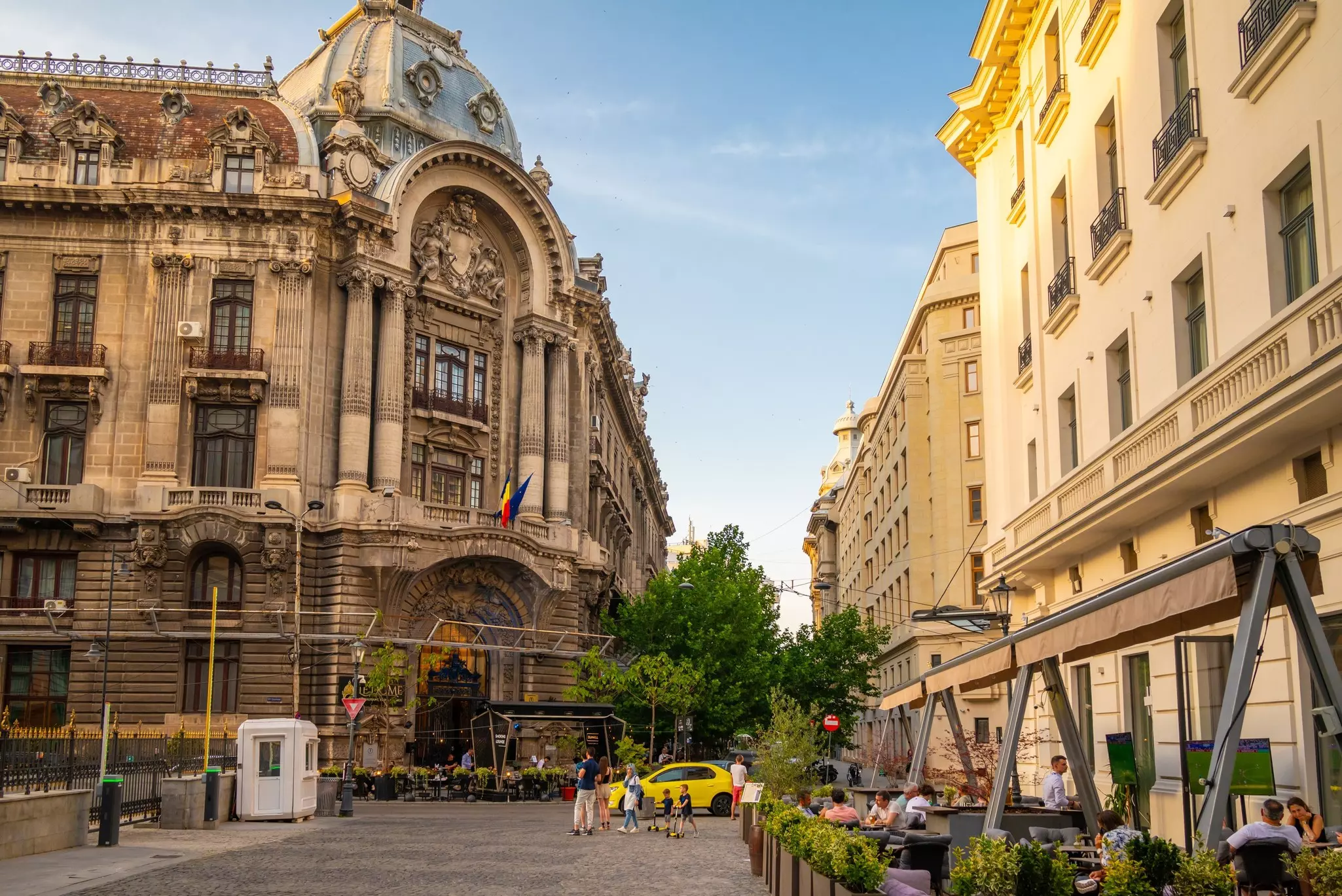 Families cross a street, while groups of people eat at outdoor cafes in front of historic buildings in downtown Bucharest.