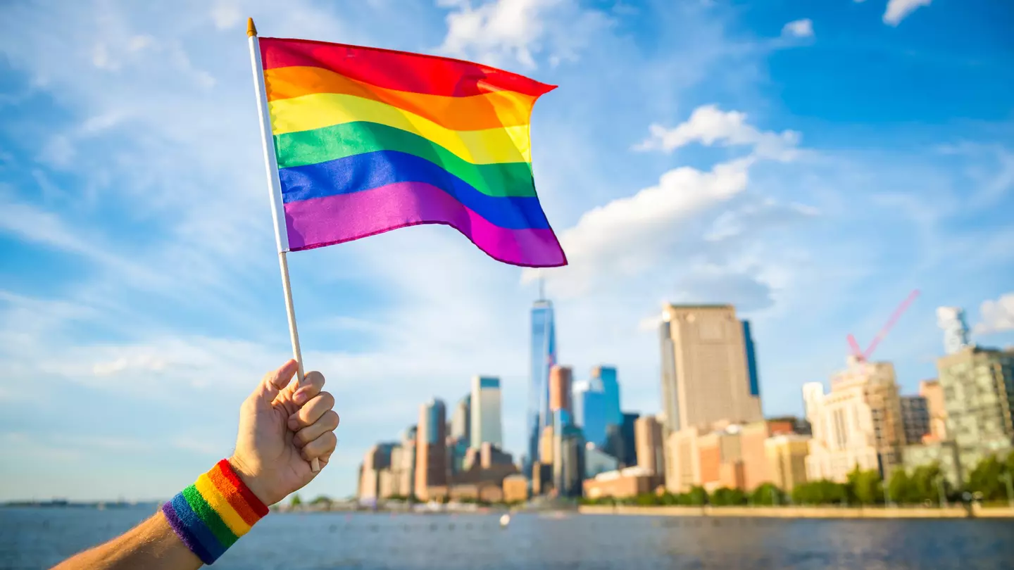 Hand holding up a colourful rainbow gay pride flag waving in the breeze with the New York City skyline in the background.