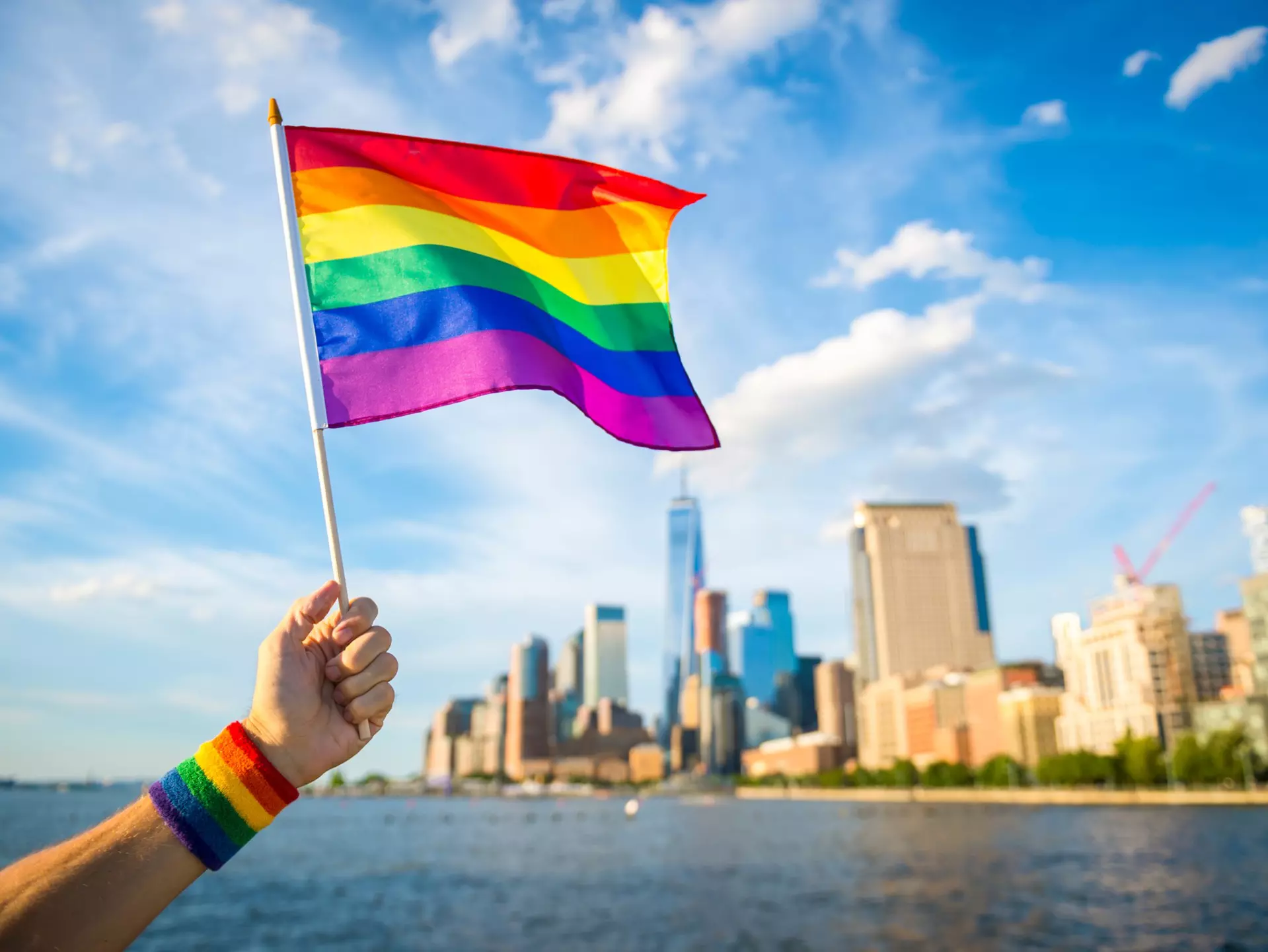Hand holding up a colourful rainbow gay pride flag waving in the breeze with the New York City skyline in the background.