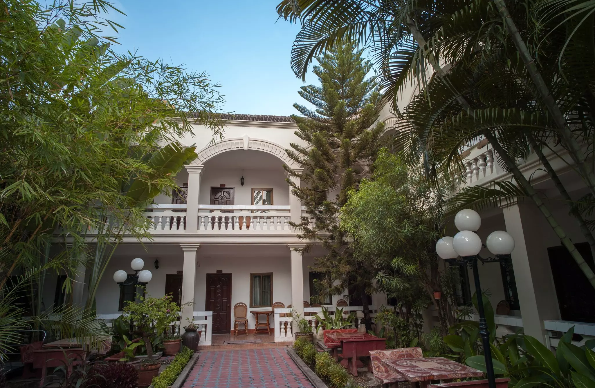 Courtyard with bamboo leading to the entrance of a white, two-story hotel with a long balcony.
