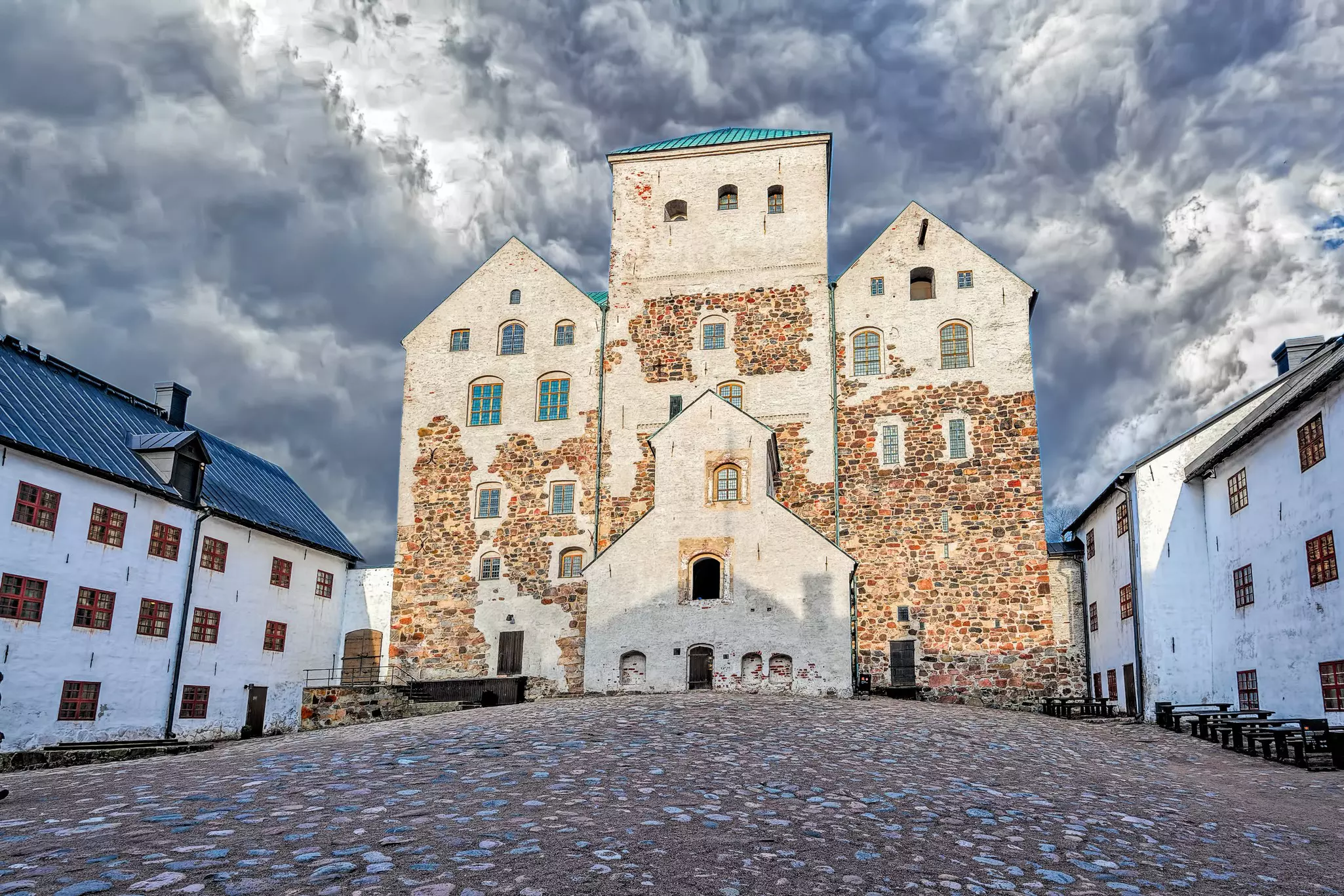 A medieval fortress with stone walls is pictured under a cloudy sky. A cobbled courtyard lies in front of the building.