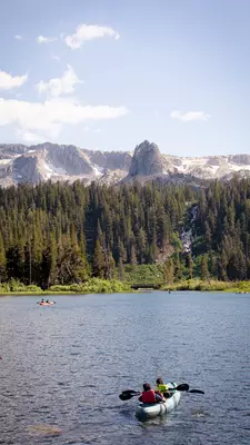 People paddle a tandem kayak in a deep-blue lake with a pine forest on the far shore; rocky peaks with patches of snow are in the background.