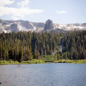 People paddle a tandem kayak in a deep-blue lake with a pine forest on the far shore; rocky peaks with patches of snow are in the background.