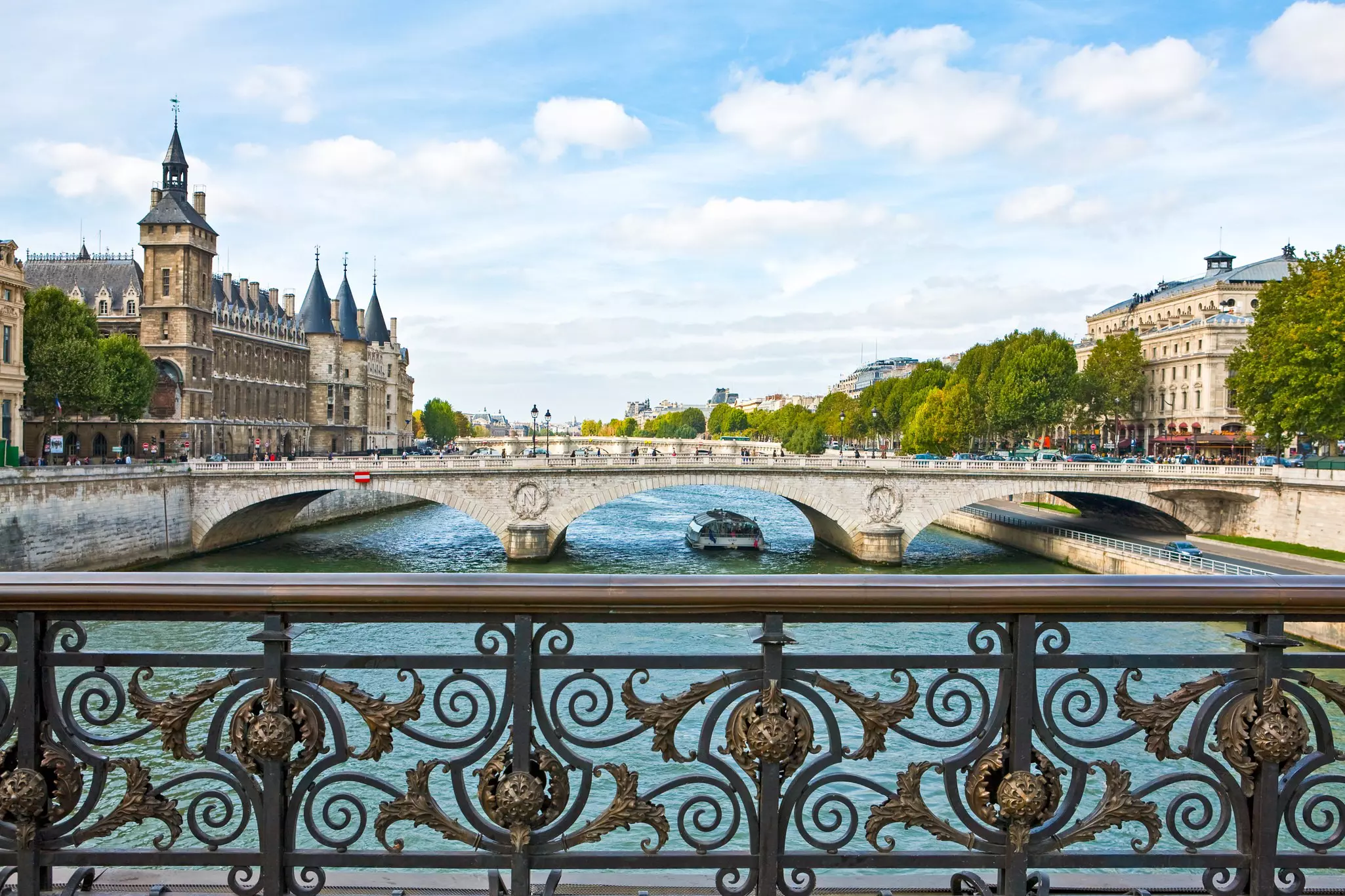 A boat passes under the archway of a bridge in Paris; the elaborate railing of another bridge is in the foreground.