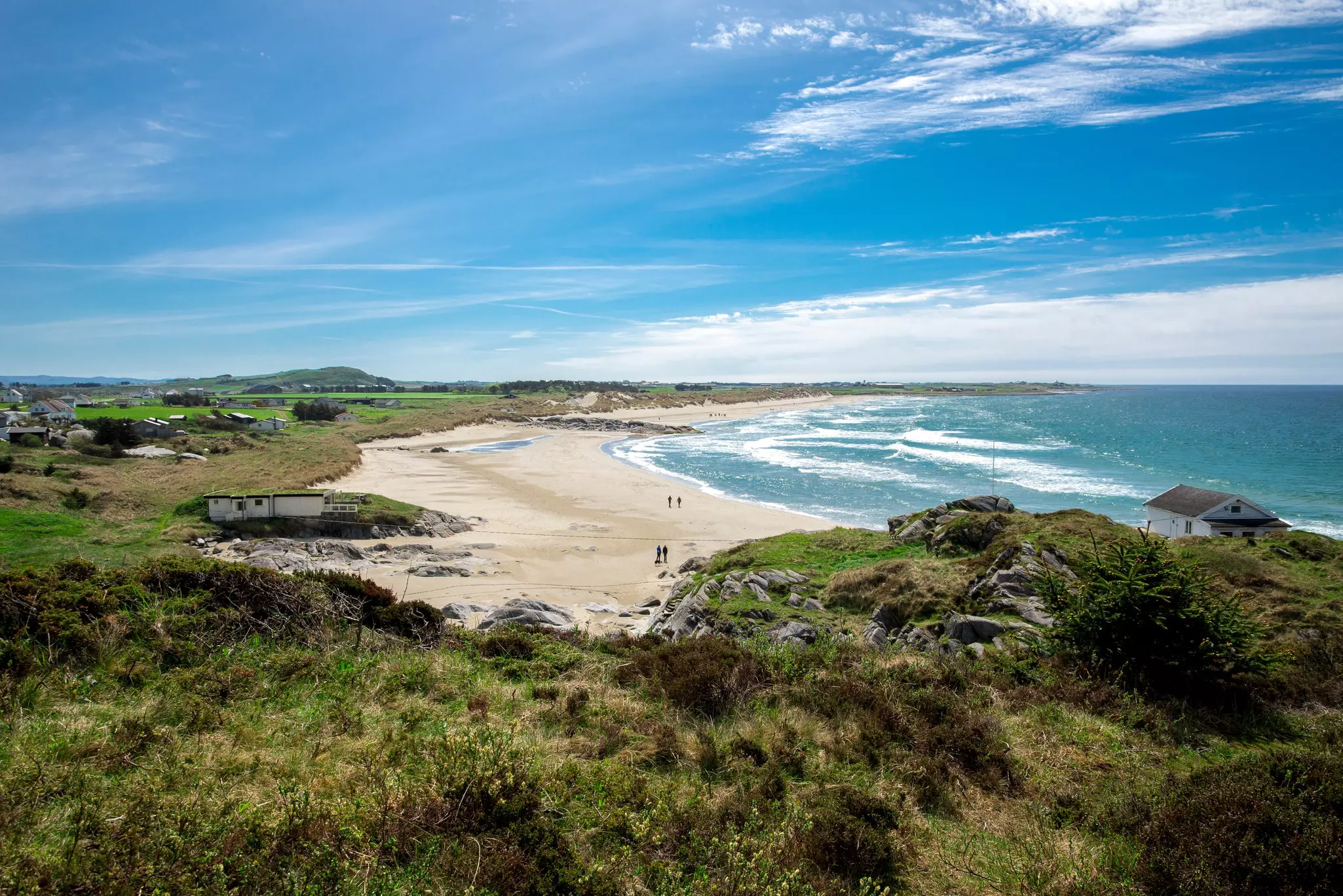 A view of a beach in Norway lapped by white waves, with dunes and grasses surrounding the sand.
