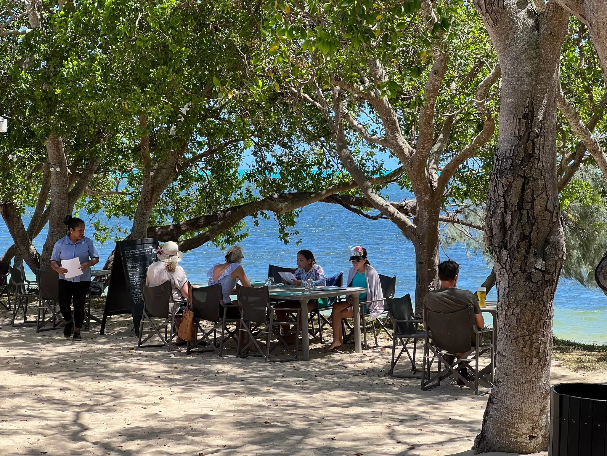 Diners at tables under trees on a sandy beach.
