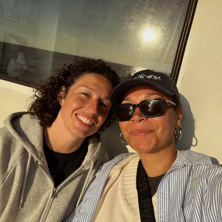 A selfie of two women smiling while sitting on a ferry on a sunny day. One woman is wearing sunglasses and a hat.