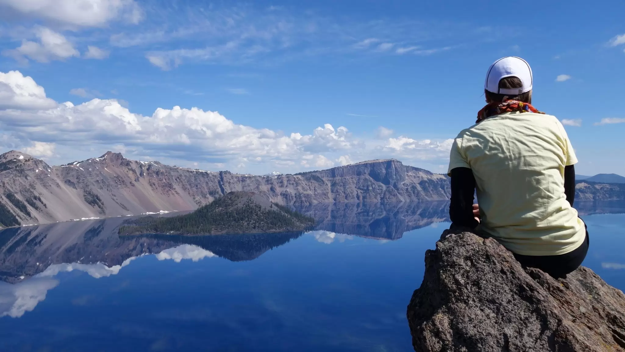 Person in a white cap sits on a rock overlooking the deep blue water and Wizard Island of Crater Lake under a bright sky