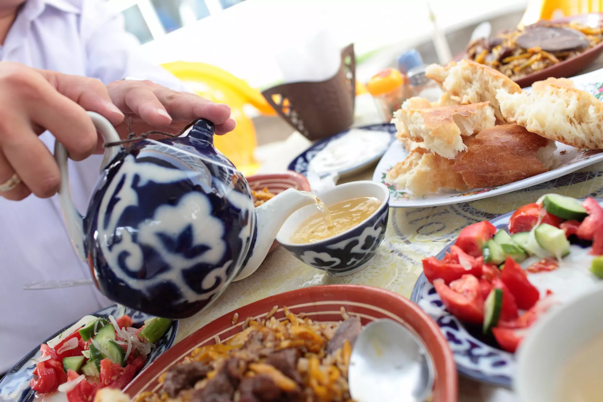 A hand pours tea from a pot at a table laden with meat and vegetable dishes in Uzbekistan.