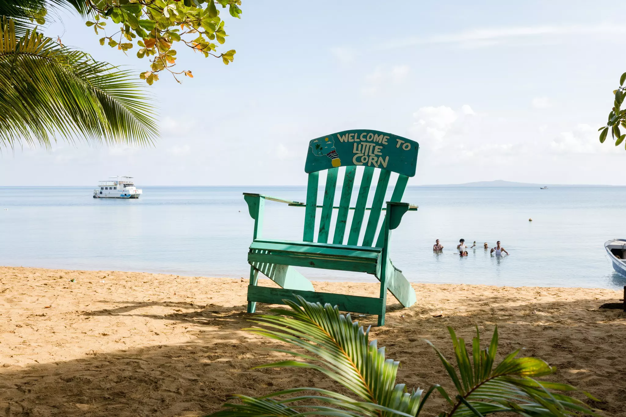 A wooden turquoise chair sits on a white sand beach and reads Welcome to Little Corn. The ocean in the background has people swimming and a large boat anchored off shore.
