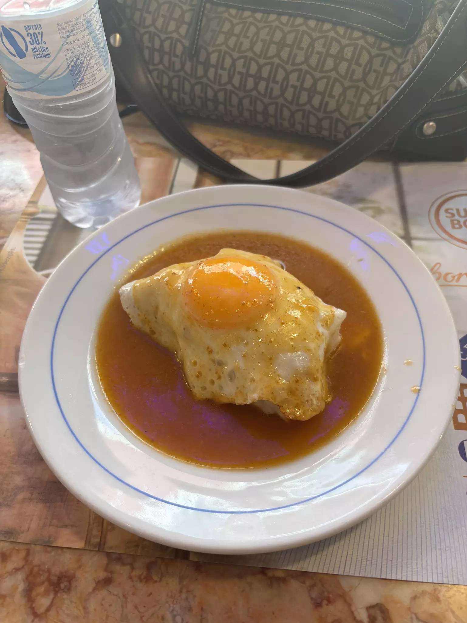 A sandwich in sauce with a fried egg on top on a white plate. The bottom halves of a water bottle and a purse are visible at the top of the photo.