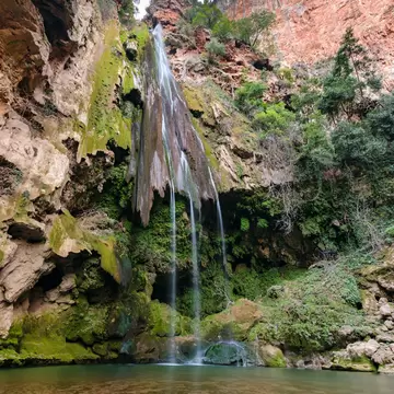 Thin waterfall streaming down a moss-covered cliff into a still pool, with red rock walls and trees above.