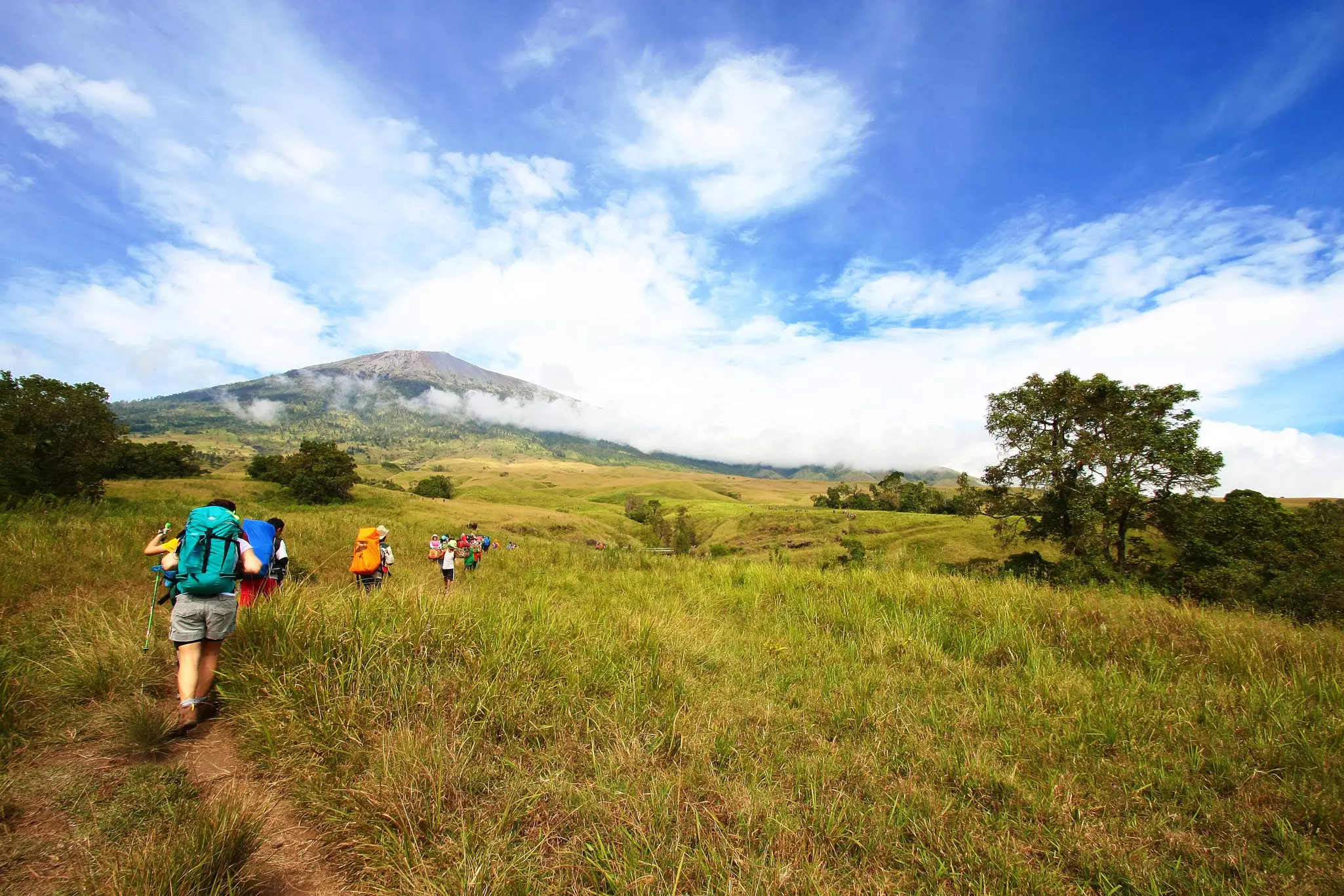 A line of hikers follow a dirt path through grassland heading up towards a volcano as low cloud rolls in.